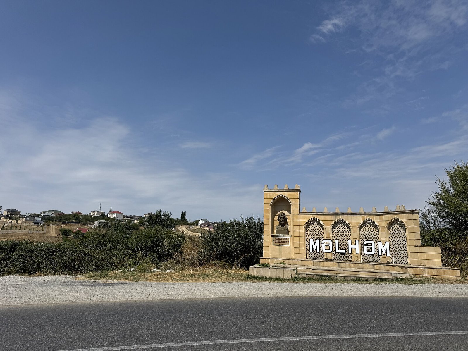 Məlhəm kəndinin girişində adı yazılmış daş abidə və yol, arxada kənd evləri, Şamaxı rayonu